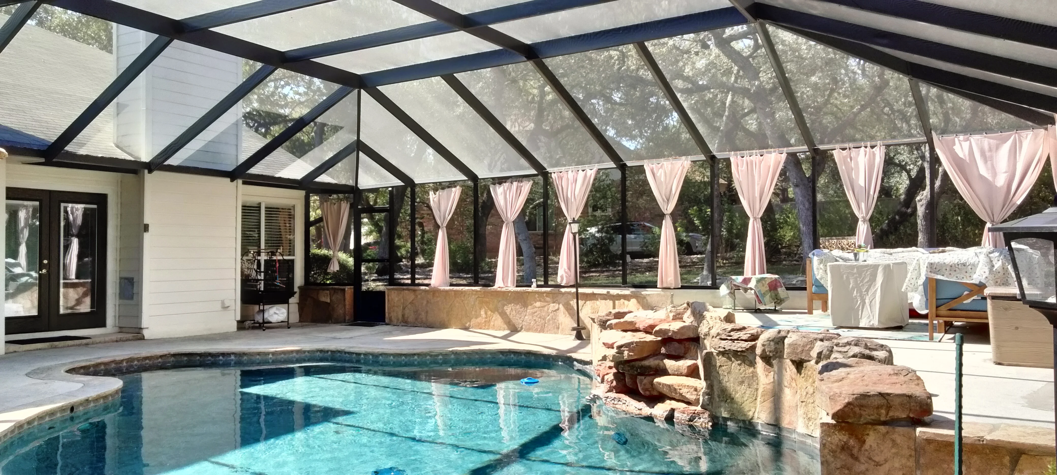 Aluminum pool enclosure with polycarbonate panels, black frame, stone water feature, and pink curtains