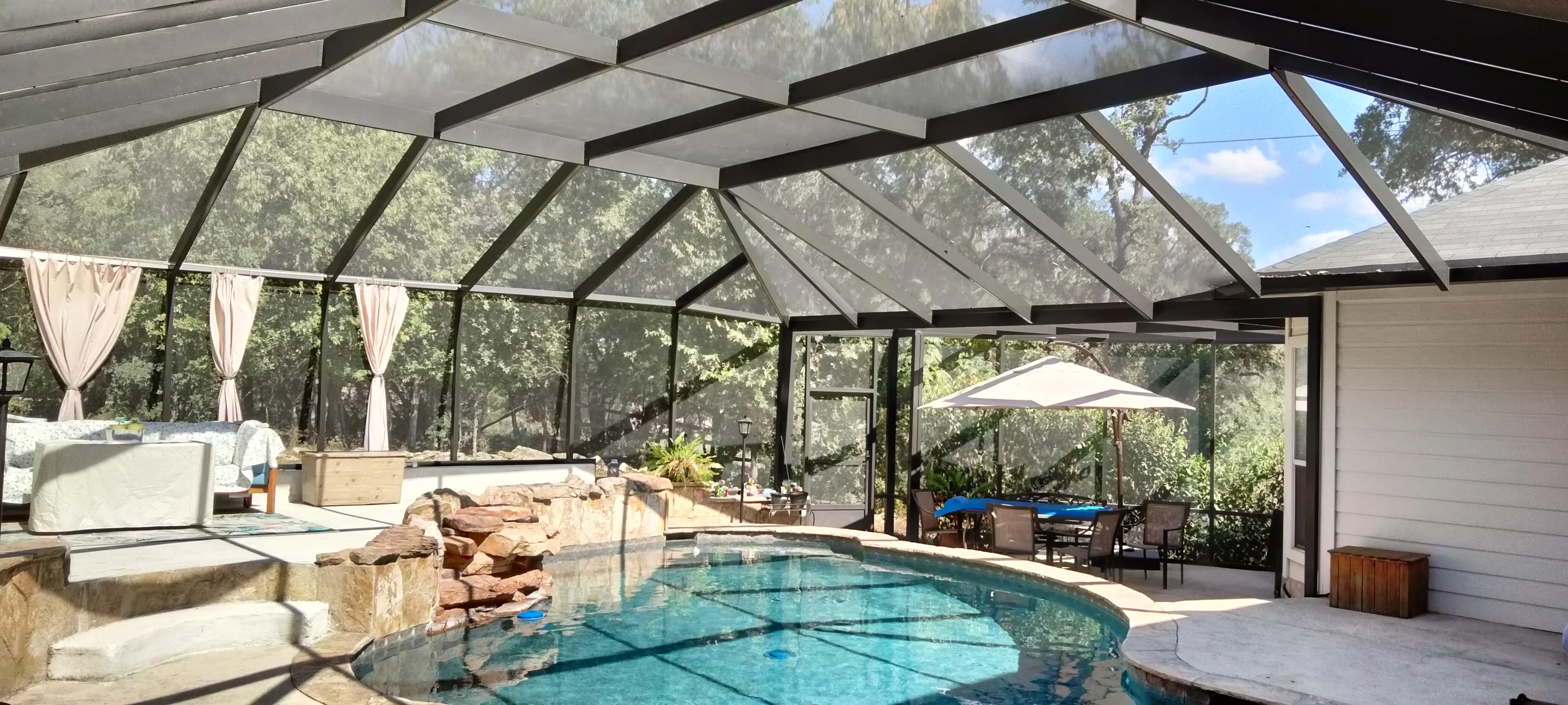 Glass pool enclosure with black aluminum frame, angled roof panels, stone waterfall feature, and outdoor furniture