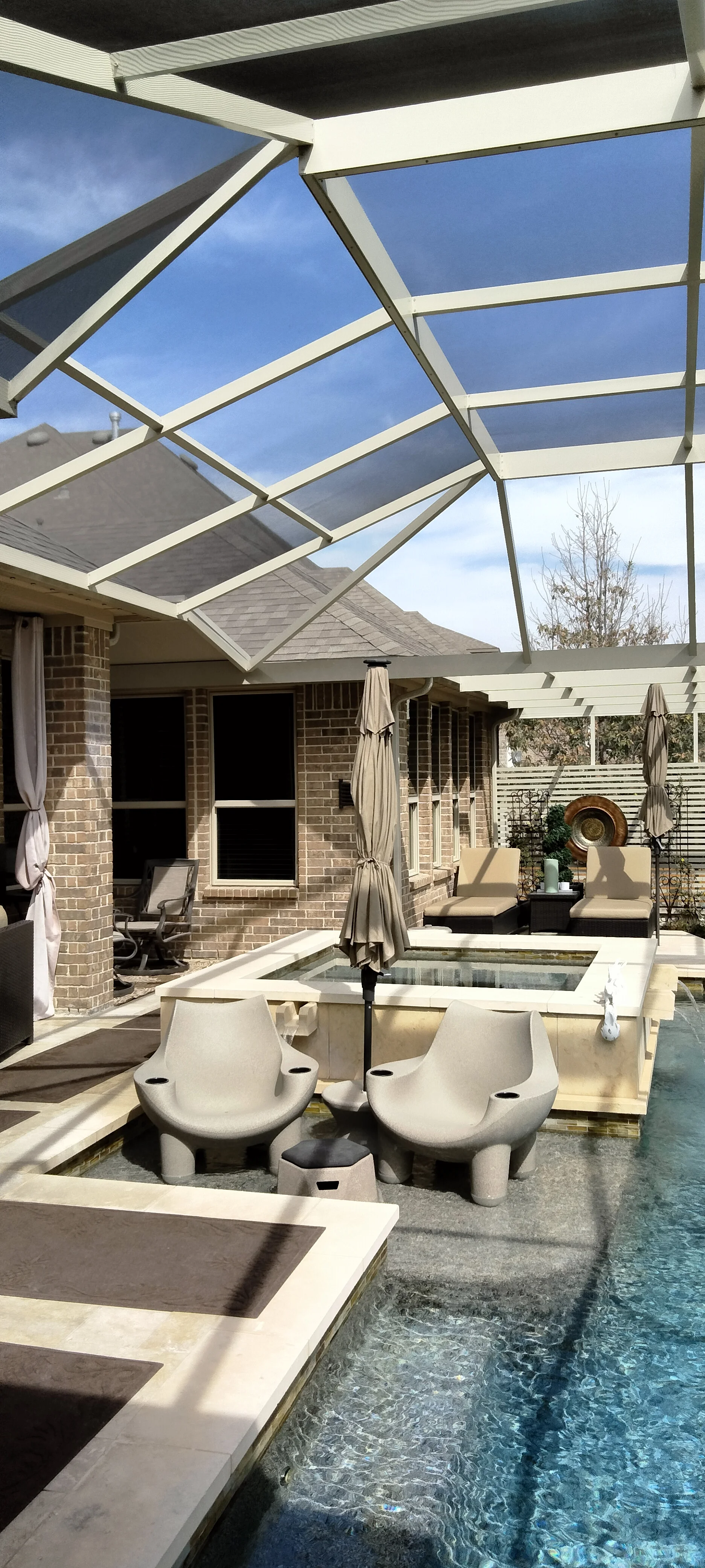 White pergola with louvered roof panels over patio with pool, brick home, and shade umbrellas