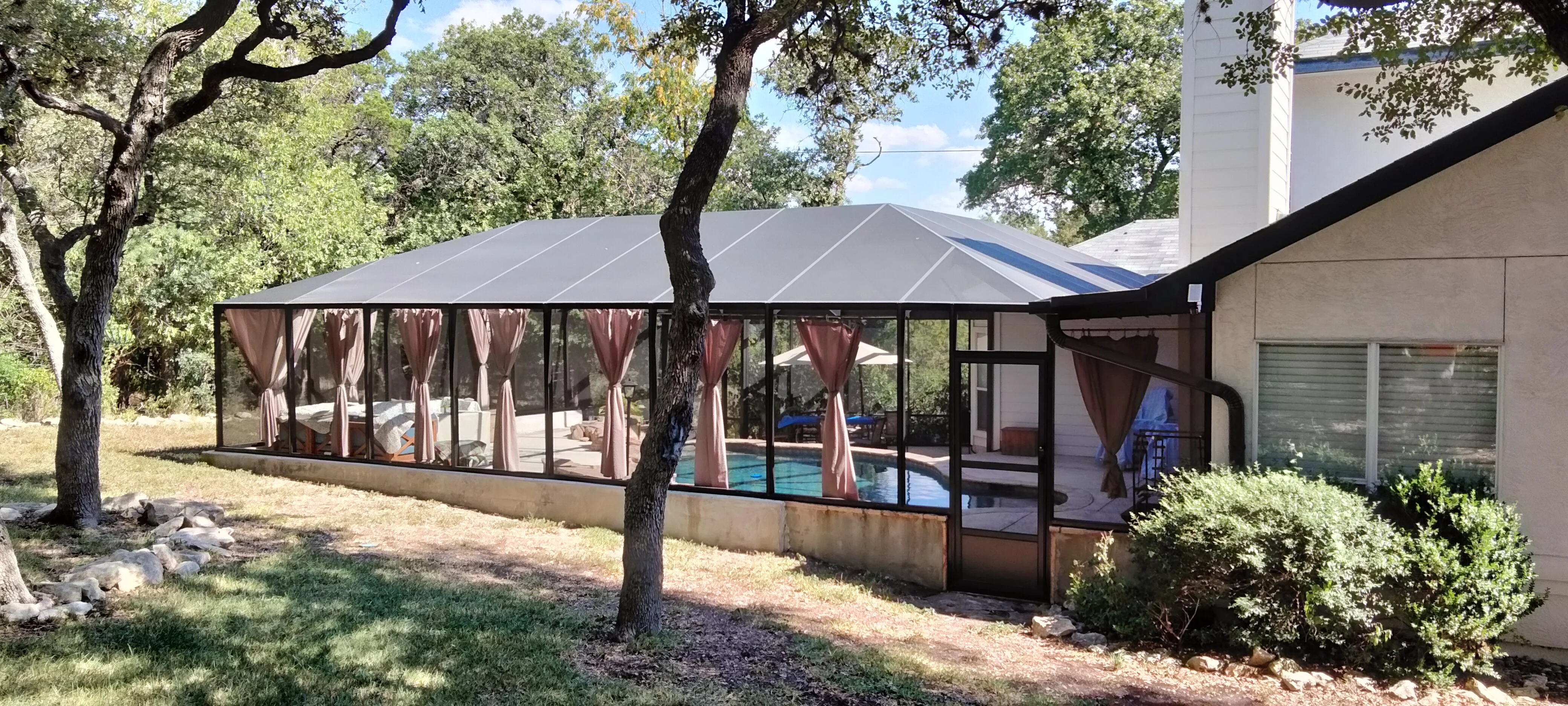Metal-framed pool enclosure with glass panels, retractable curtains, and standing seam roof
